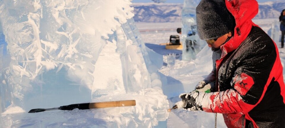 Man in red jacket chisels a block of ice into a sculpture outdoors in winter.