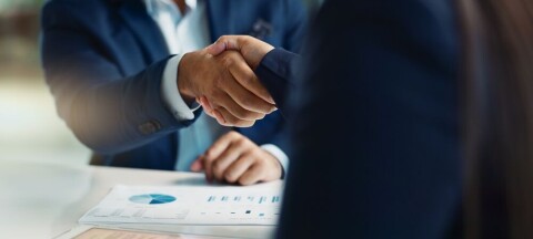 Two people in business suits shaking hands over documents with charts on a desk